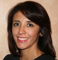 The image shows a smiling woman with dark hair, wearing a black top, posing for a portrait against a neutral background.