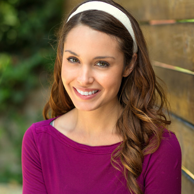 The image features a woman with long brown hair wearing a purple top and a headband, smiling at the camera against a wooden fence background.