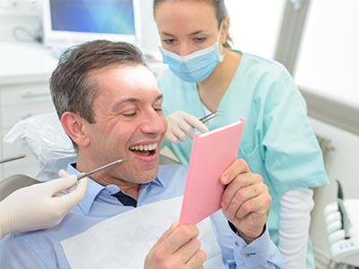 The image shows a man sitting in a dental chair with his mouth open, holding up a pink card, while a female dentist stands behind him, smiling and looking at the card.