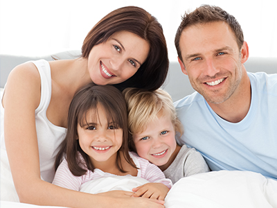 A family of four, including two adults and two children, posing together on a bed with smiles.