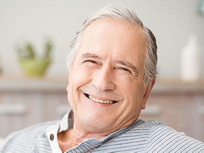 The image shows a smiling older man with grey hair, wearing a dark shirt, seated comfortably in a relaxed posture.