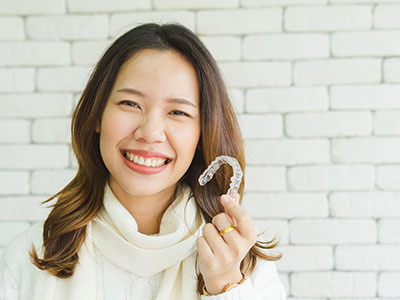 A woman with a smile holds up a dental retainer, standing against a brick wall.